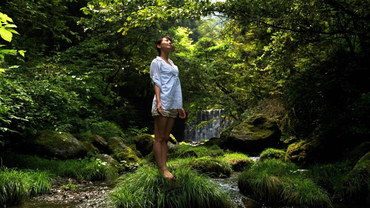 A woman standing barefoot on a small grassy patch in the middle of a shaded, tree lined creek.