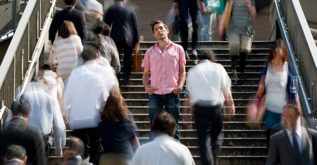 A man standing on a staircase as people move around him, he's the only one not blurred by motion and he's facing the camera while everyone else walks the other way.