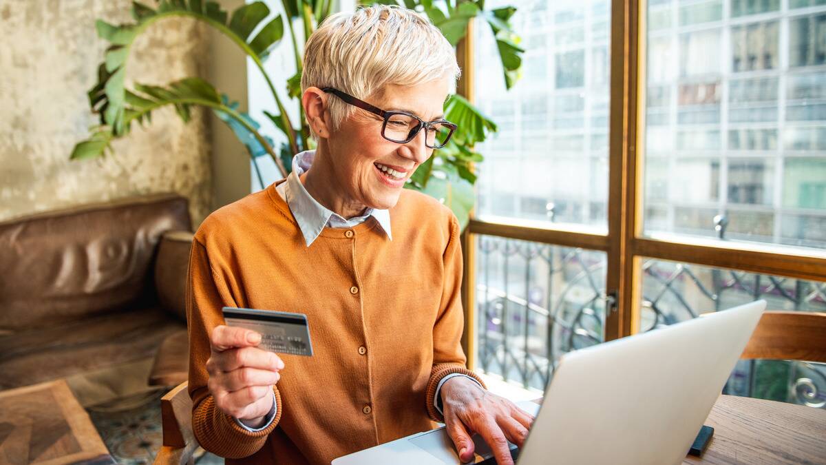 A woman sitting t her computer, smiling, holding up her credit card as she pays for something online.