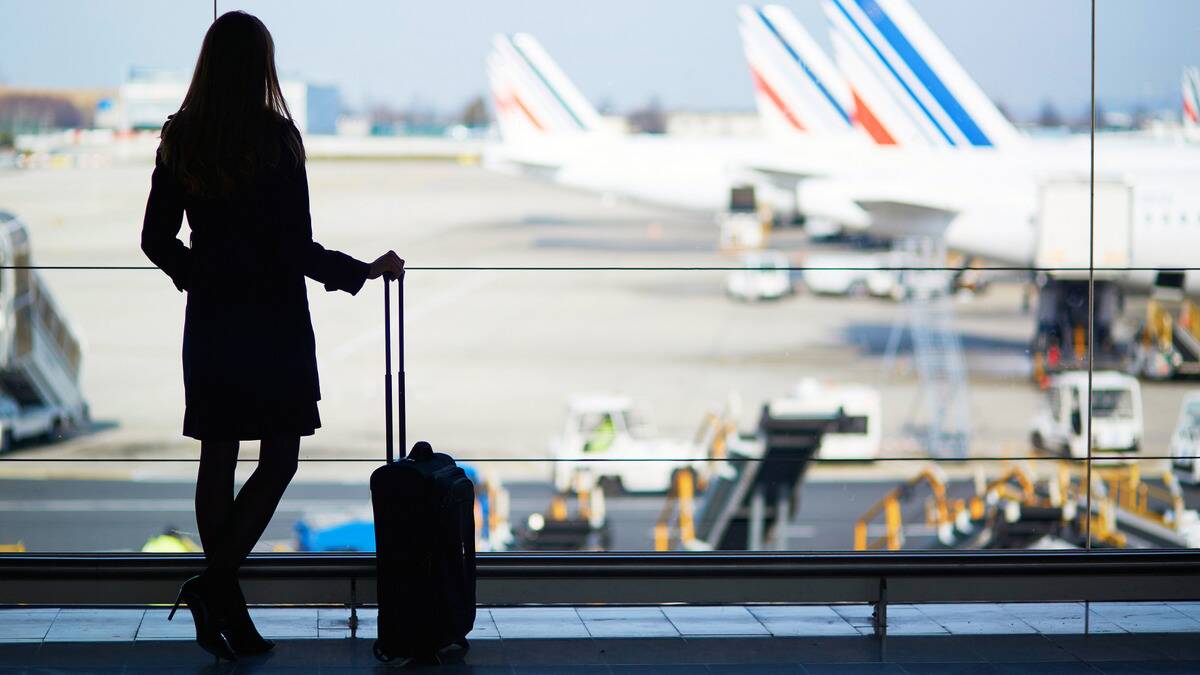 A silhouette of a woman and her small carry-on suitcase standing in front of a large airport window, overlooking the planes.