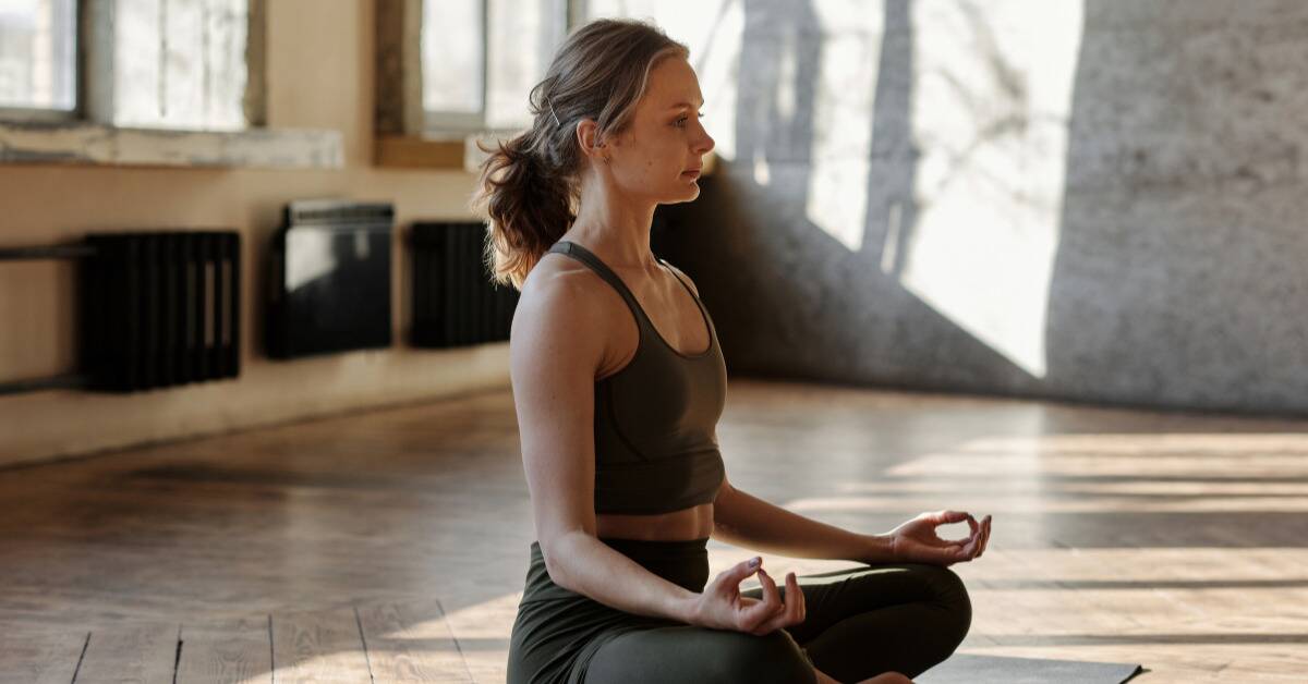 A woman meditating on a yoga mat in the center of an empty room.