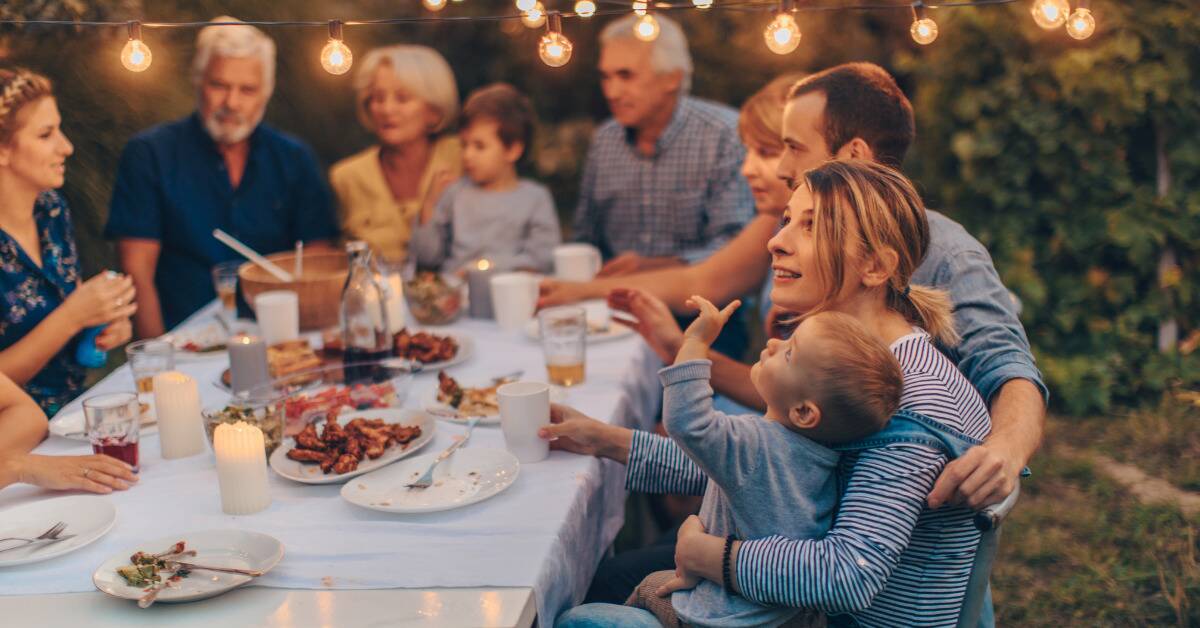A large family gathering around a table, the focus on a mom holding her young son in her arms as he points toward one of the hanging lights.