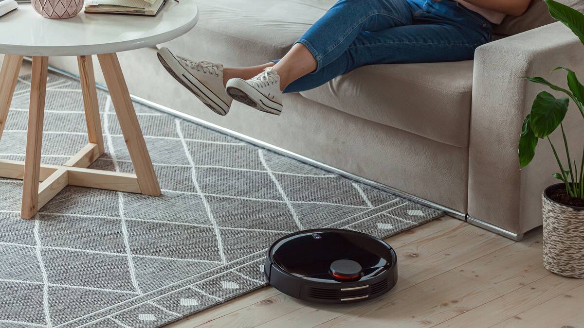 A robot vacuum about to clean an area rug, a girl sitting on the nearby couch lifting her legs so it can get underneath her.