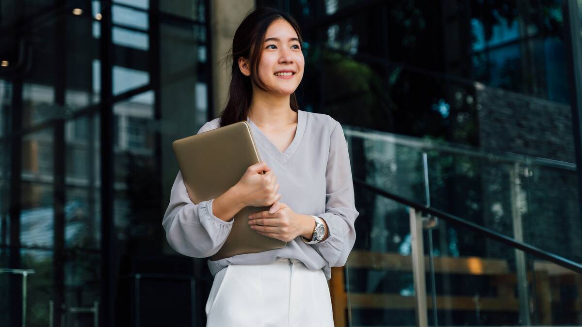 Ayoung professional woman walking out of an office building, holding her laptop and smiling.