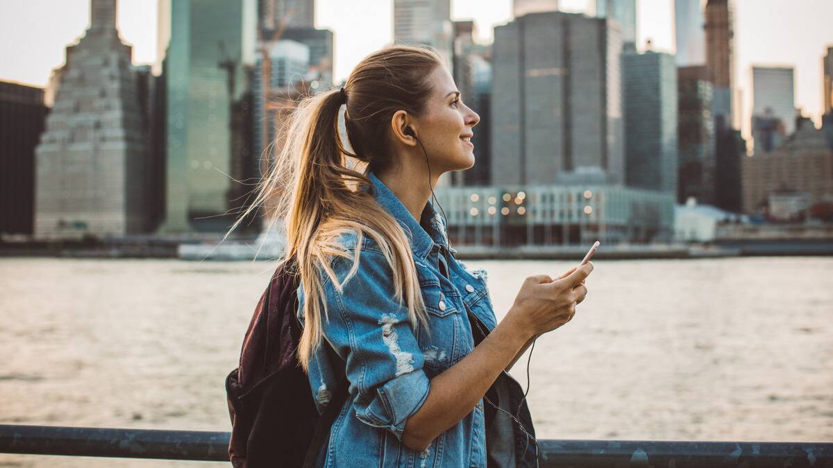 A woman walking a path along a large river next to a city, smiling as she listens to music through wired earbuds that are connected to the phone she's holding.