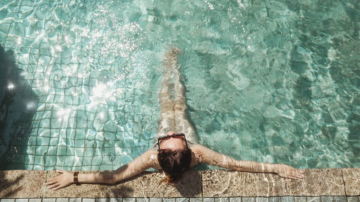 An aerial shot of a woman sitting in a pool, her arms up on a tiled ledge that's also underwater.