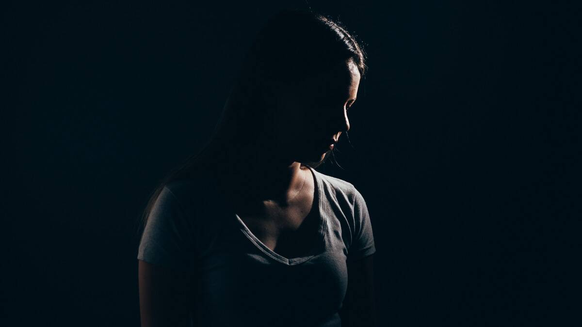 A woman standing in near darkness, looking off to the side, a small light illuminating just part of her face and shoulder.