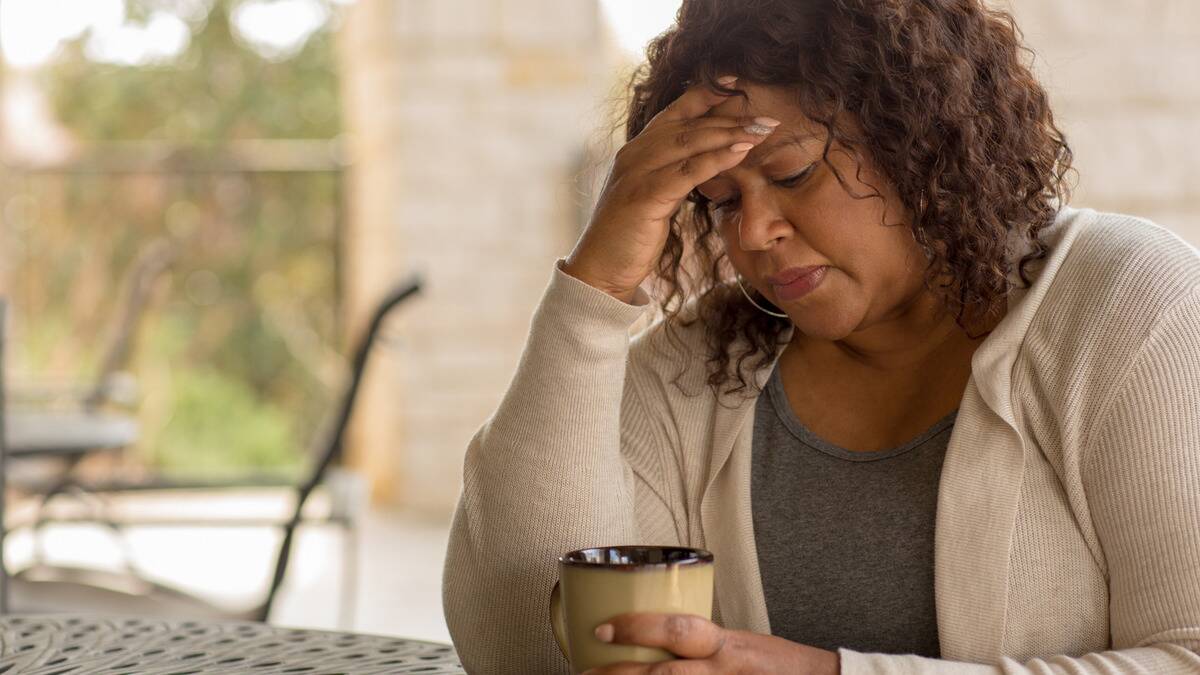 A woman sitting at a table, looking down into her mug, a hand on her forehead.