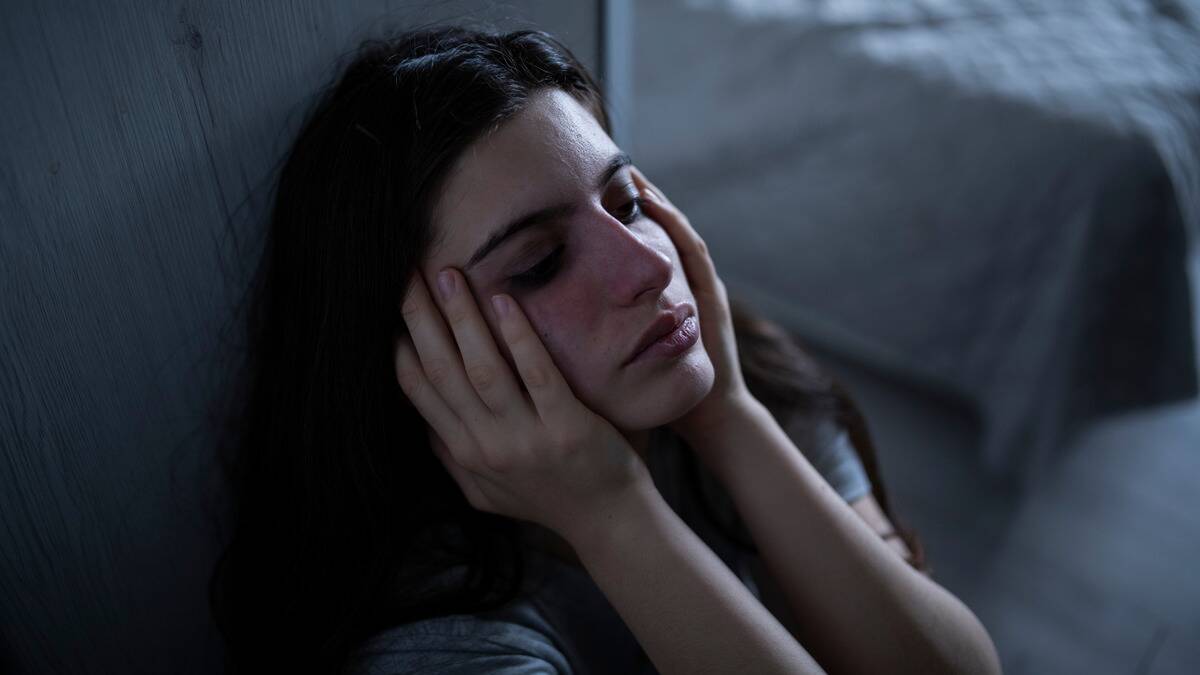 A woman sitting on the ground, back against a wall, both hands on the side of her face, looking stressed.