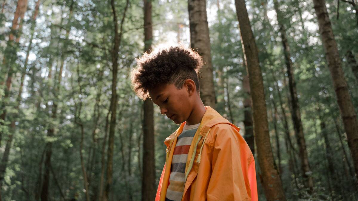 A young boy standing in a forest, looking down at his shoes.