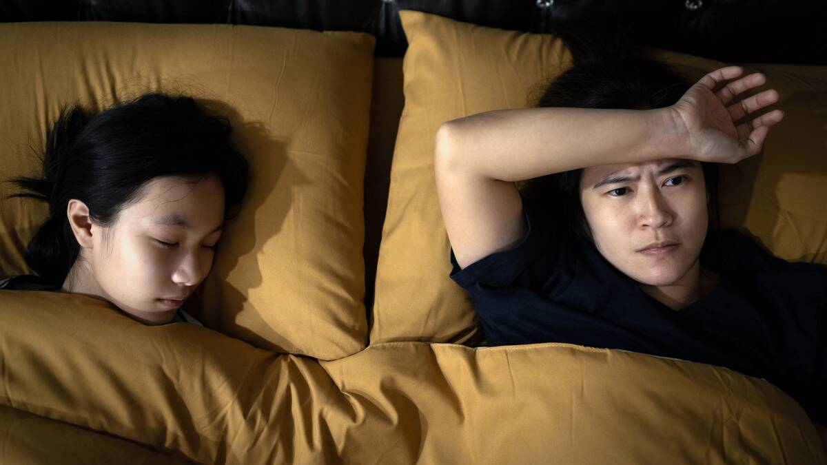 A couple in bed next to one another, the woman sleeping, the man looking anxious, arm on his forehead.