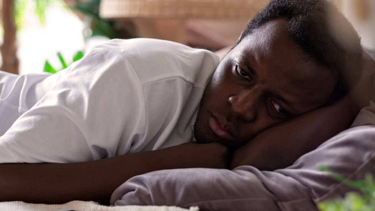 A man laying on his side in bed, arms under his head, looking stressed.