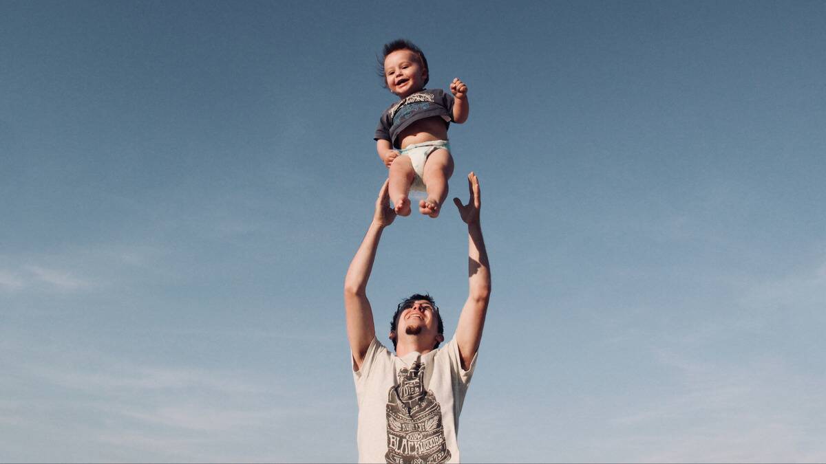 A man standing in front of a clear blue sky, tossing his baby gently into the air, both smiling.