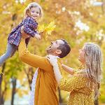 A father holding his young daughter up in the air, the mother behind him, the daughter smiling and holding a bundle of autumn leaves.