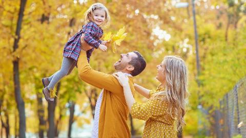 A father holding his young daughter up in the air, the mother behind him, the daughter smiling and holding a bundle of autumn leaves.