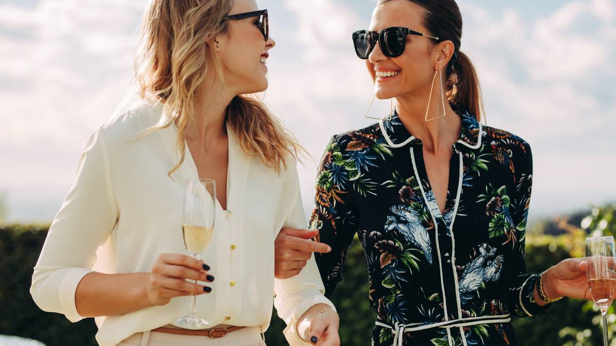 Two stylish friends smiling as they walk and chat outside, arms linked, holding glasses of champagne.