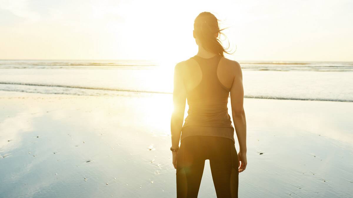 A woman standing powerfully on the beach, facing the water, the sun rising before her.