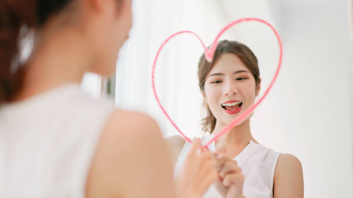 A woman drawing a heart around her face in the mirror with lipstick.