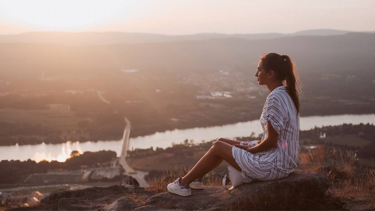 A woman sitting on a hill above a city, the sun shining on her.