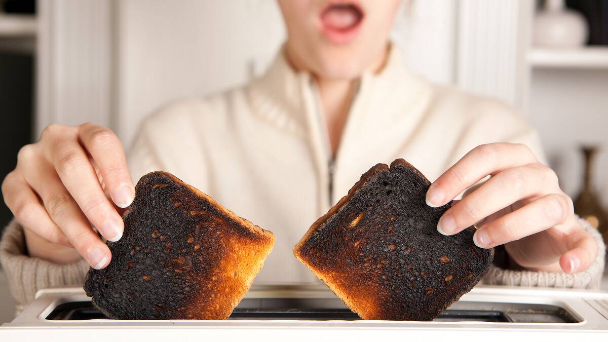 A woman pulling out two pieces of blackened, burnt toast out of the toaster, her face seeming shocked in the background.