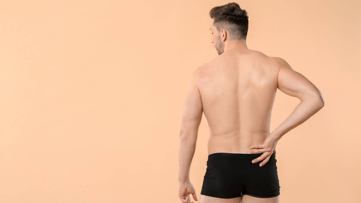 A man against a skin-toned backdrop, back to the camera, wearing black boxer briefs.
