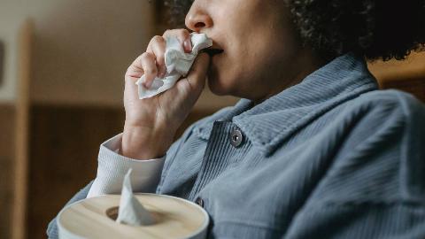 A close shot of a woman holding a box of tissues in one hand, a balled up tissue in the other up to her nose and mouth.