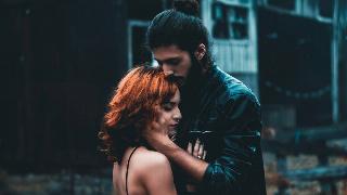 A couple standing outside, seeming in heavy shadow, the woman with her head and hands against the man's chest, the man holding her jaw and pressing his lips to the crown of her head.