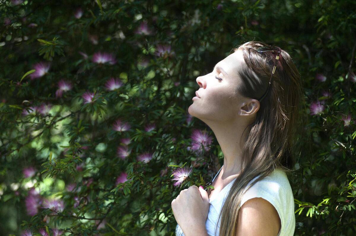 A woman standing outside amidst a flowering tree ot hedge, eyes closed, head looking up, hand to her chest.
