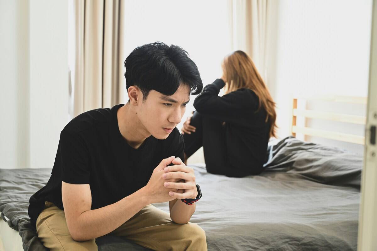 A couple sitting on opposite sides of the bed, the man with his elbows on his knees, leaned forward, looking contemplative. The woman is facing away from the camera, but we can see her knees drawn to her chest and a hand on her head.