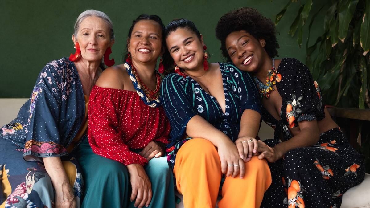 A group of four women, all in bright clothing, sitting next to one another and smiling as they lean into each other.