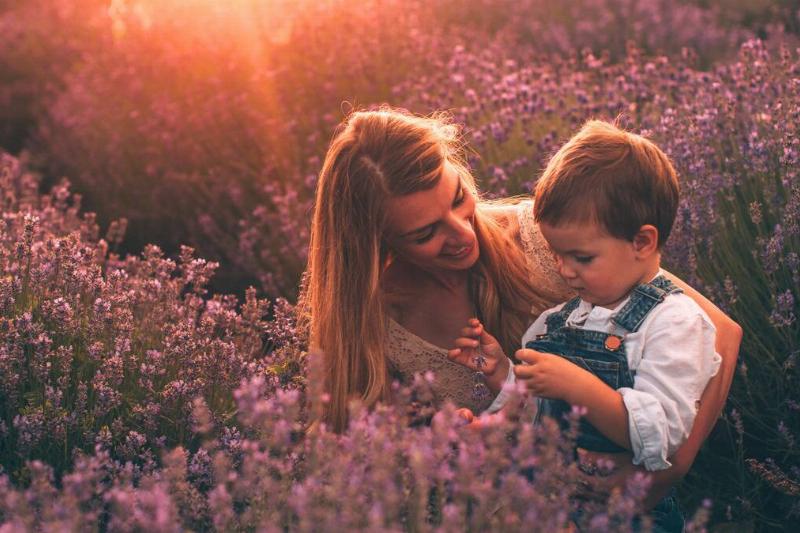 A woman kneeling in a field of small purple flowers with her young son, the sun shining on them.