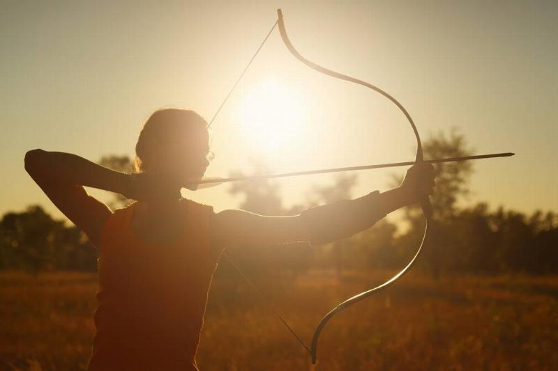 A heavily silhouetted image of a woman drawing back a traditional-style bow, the sun behind her.