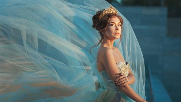 A woman in a crown and fancy ballgown, the outer layer of tule billowing in the wind, looking off to the side pensively.