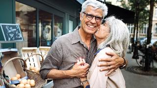 A couple standing outside, arms around one another, the man smiling as the woman kisses his cheek.