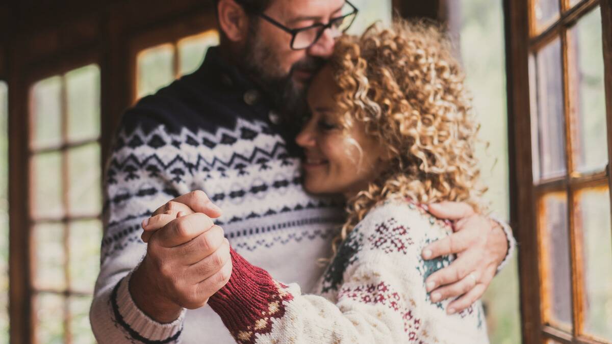 A couple slow dancing inside in Christmas sweaters, the focus on their hands together.