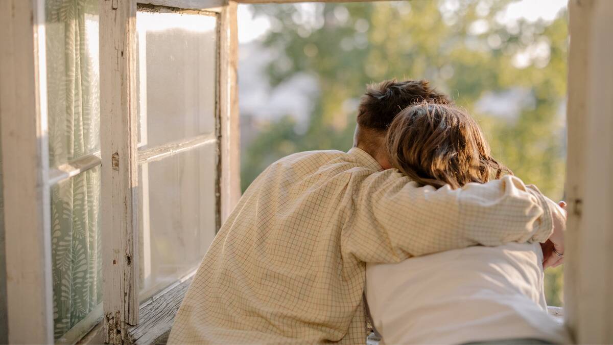 A couple where the man has his arm around his girlfriend, facing away from the camera, half leaning out a window so they can look outside.