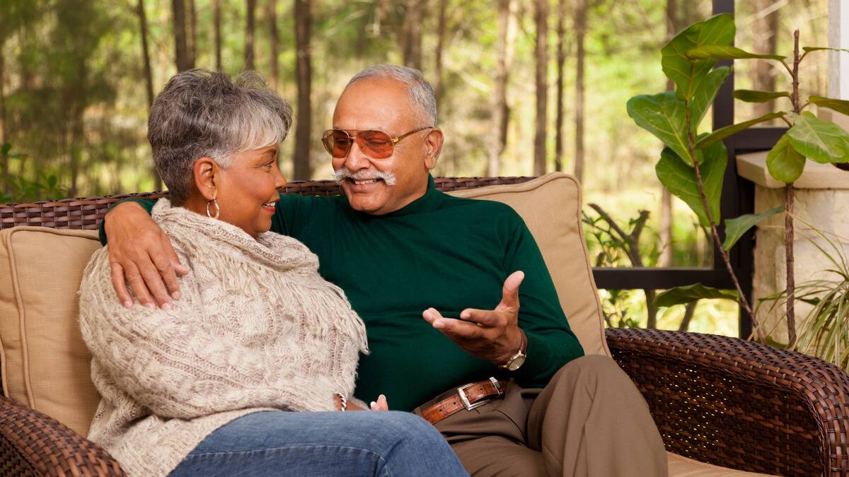 A couple sitting on a loveseat outdoors, the man with his arm around the woman, both smiling at they chat.