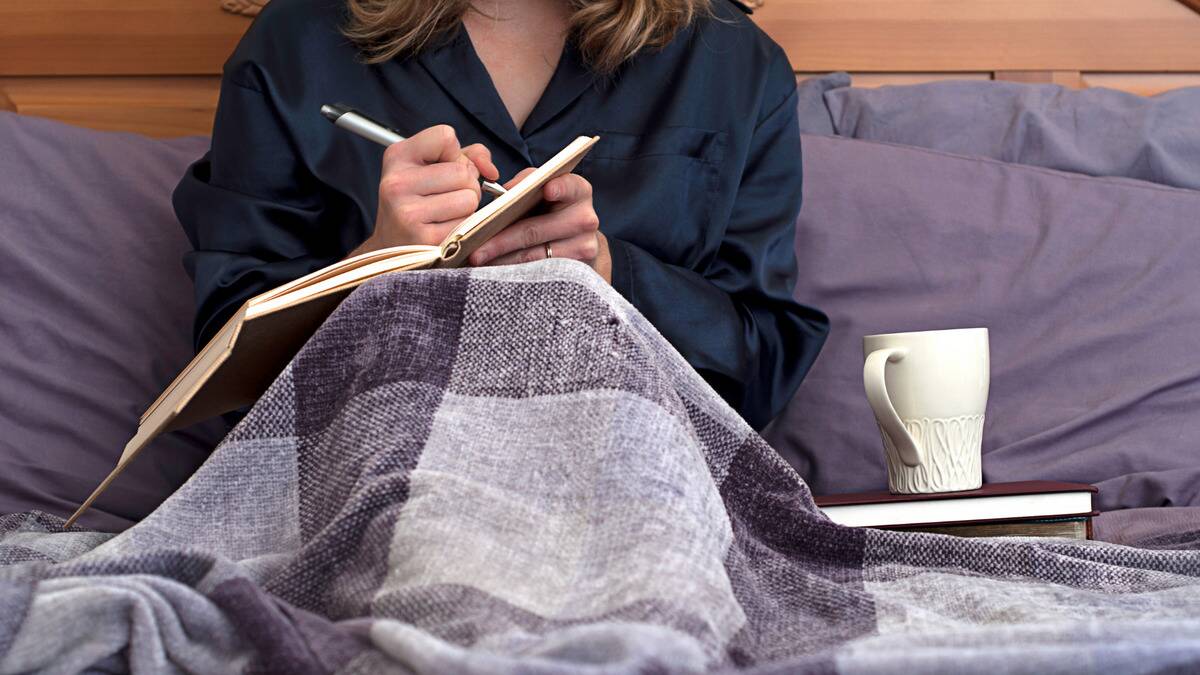 A woman sitting in bed, blanket over her legs, writing in a journal against her knees.