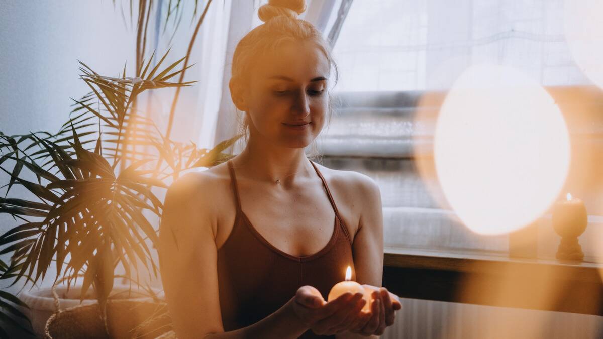 A woman sitting by a window in the sun holding a small, round, lit candle, smiling peacefully.