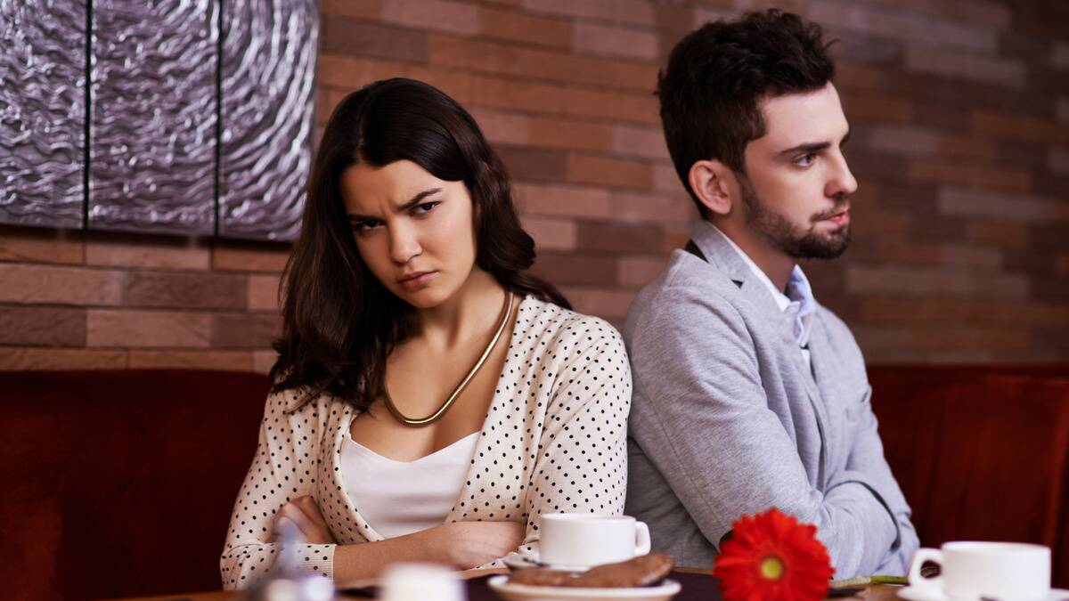 A couple sitting side by side in a restaurant, back to bad, both with their arms crossed and looking frustrated.