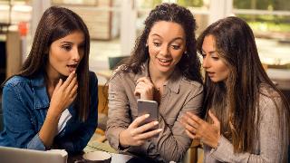 A woman showing her two friends something on her phone, all looking stunned.