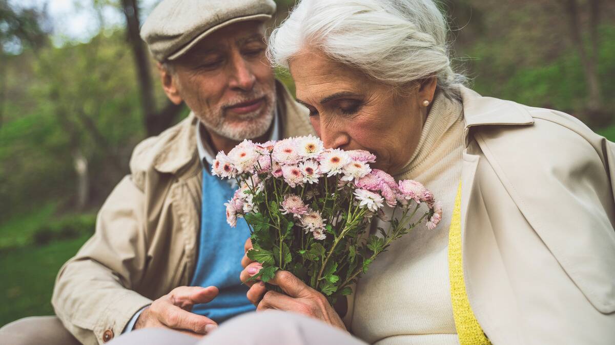 A close shot of a couple sitting together, the woman holding a bouquet of flowers and smelling them with her eyes closed, the man watching her.