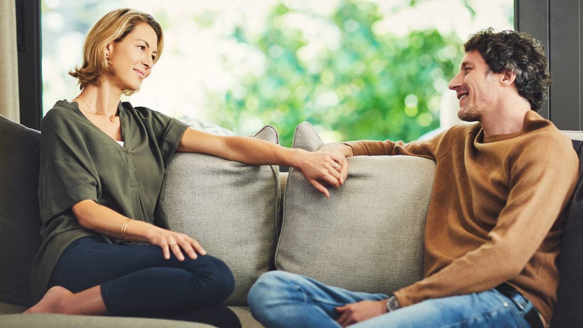 A couple amicably sitting on opposite ends of a couch, facing each other, arms resting on the back of the couch, smiling as they chat.