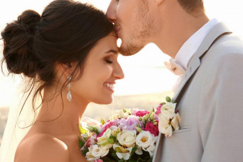 A close shot of a couple at their wedding, the husband kissing his wife on the forehead, the wife with her eyes closed and smiling, holding her bouquet between their chests.