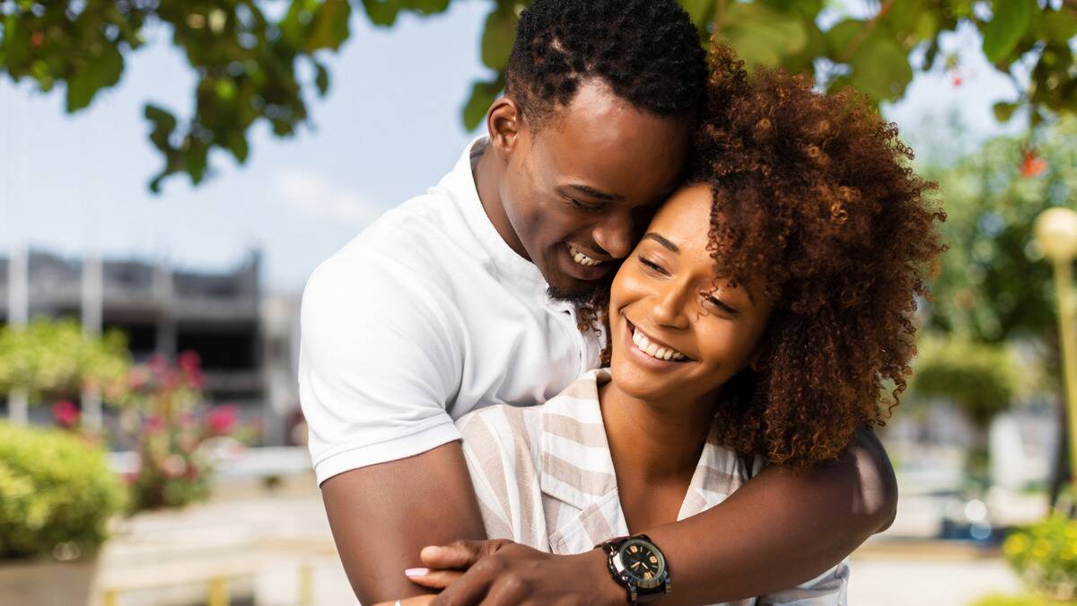 A couple standing together outside, both smiling, the man behind the woman and hugging her from behind.