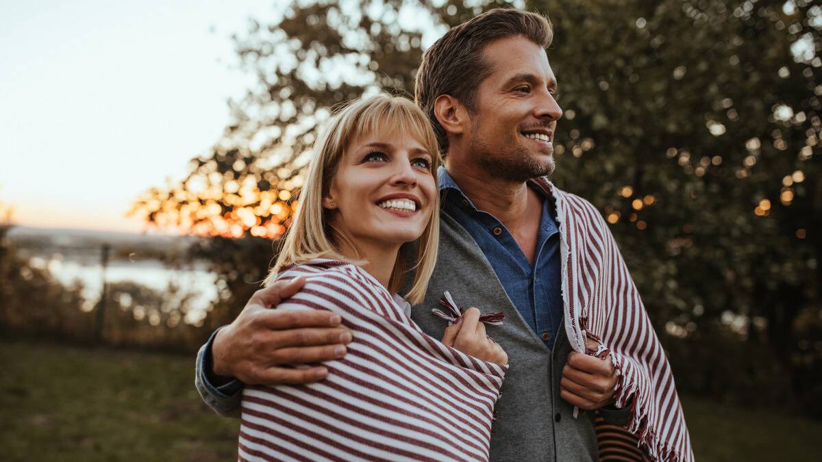 A couple walking outside, both smiling, a blanket wrapped around both of their shoulders.