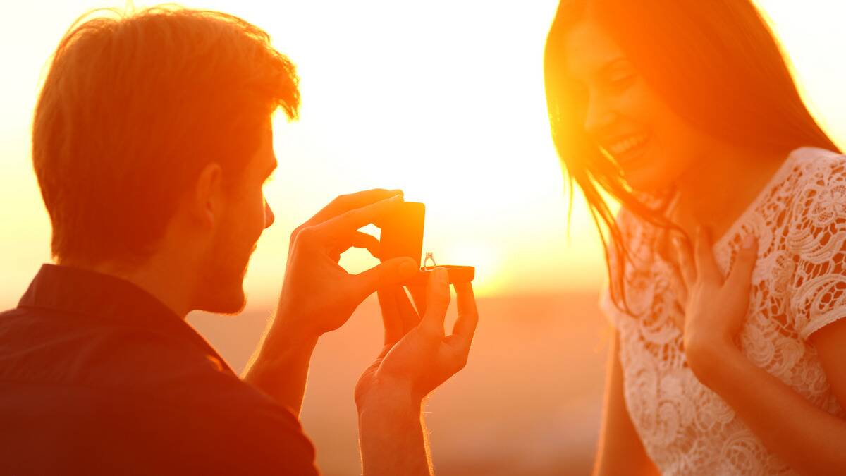 A close shot of a man proposing to his girlfriend, the sun setting behind them, washing them in an orange glow.