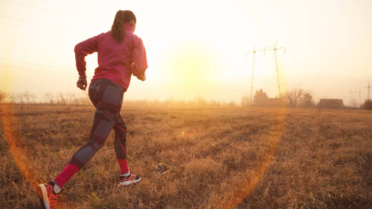 A woman in running gear running through a field, the sun shining behind her.