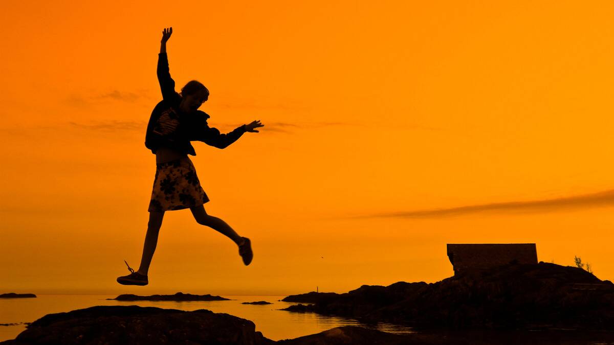 The silhouette of a woman jumping in the air gleefully at the waterfront, an orange sky behind her.