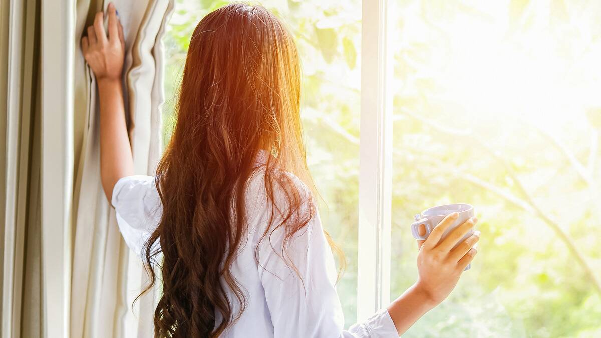 A woman drawing open a curtain to look out the window, a mug in her other hand.